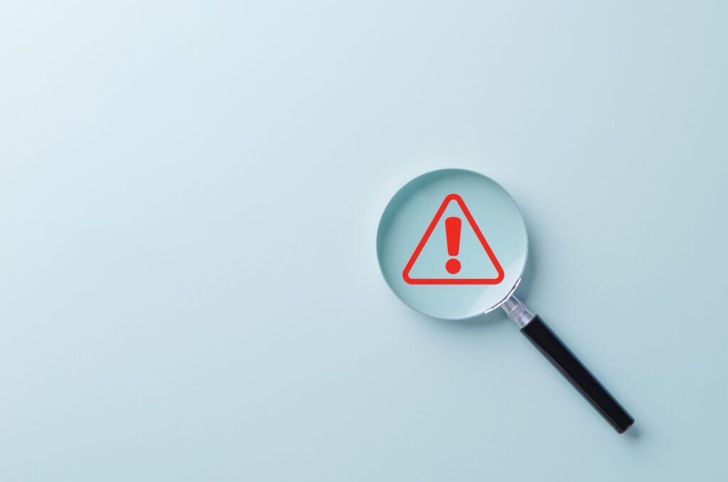A red warning symbol under magnifying glass, symbolizing fraud detection.